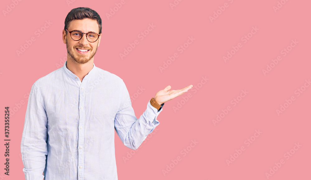 Handsome young man with bear wearing elegant business shirt and glasses smiling cheerful presenting and pointing with palm of hand looking at the camera.