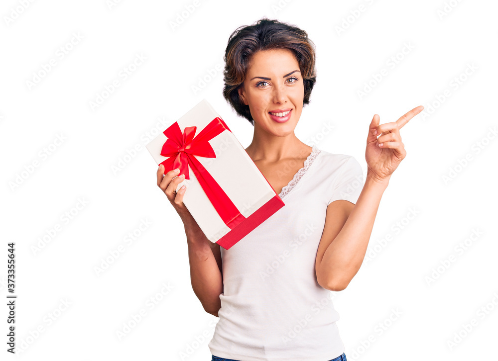 Young beautiful woman holding gift smiling happy pointing with hand and finger to the side Young beautiful woman holding gift smiling happy pointing with hand and finger to the side