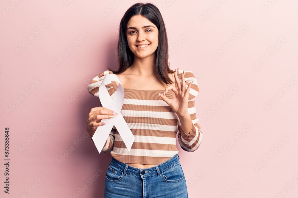 Young beautiful girl holding white cancer ribbon doing ok sign with fingers, smiling friendly gesturing excellent symbol Young beautiful girl holding white cancer ribbon doing ok sign with fingers, smiling friendly gesturing excellent symbol