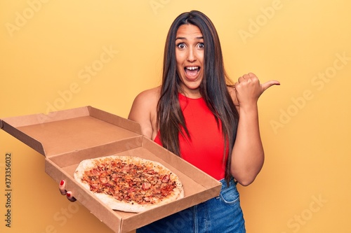 Young beautiful brunette woman holding delivery box with italian pizza pointing thumb up to the side smiling happy with open mouth