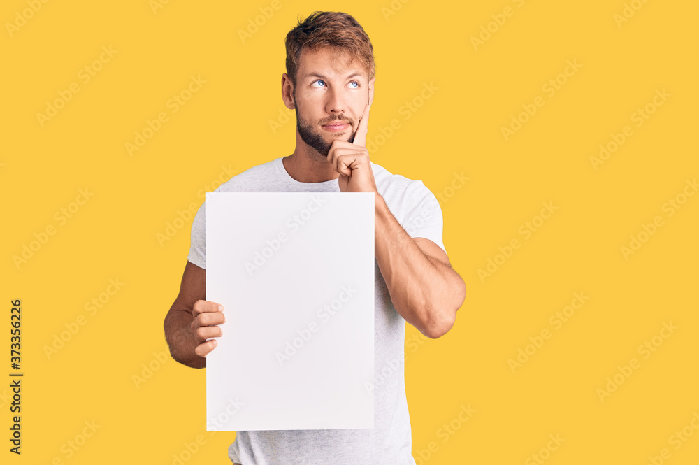 Young caucasian man holding blank empty banner serious face thinking about question with hand on chin, thoughtful about confusing idea Young caucasian man holding blank empty banner serious face thinking about question with hand on chin, thoughtful about confusing idea