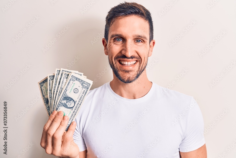 Young handsome man holding bunch of dollars banknotes over isolated white background looking positive and happy standing and smiling with a confident smile showing teeth Young handsome man holding bunch of dollars banknotes over isolated white background looking positive and happy standing and smiling with a confident smile showing teeth