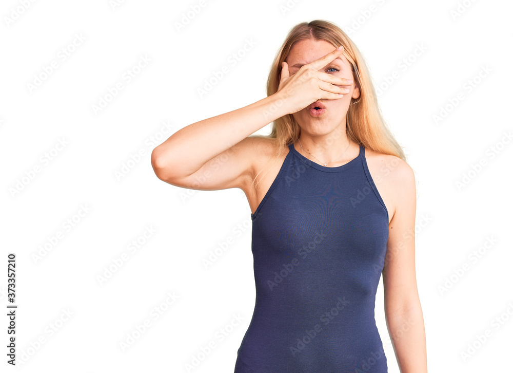 Young beautiful blonde woman wearing casual dress peeking in shock covering face and eyes with hand, looking through fingers with embarrassed expression. Young beautiful blonde woman wearing casual dress peeking in shock covering face and eyes with hand, looking through fingers with embarrassed expression.