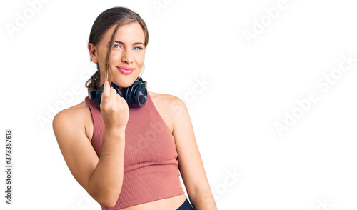 Beautiful caucasian young woman wearing gym clothes and using headphones beckoning come here gesture with hand inviting welcoming happy and smiling