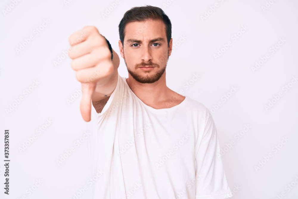 Young handsome man wearing casual white tshirt looking unhappy and angry showing rejection and negative with thumbs down gesture. bad expression. Young handsome man wearing casual white tshirt looking unhappy and angry showing rejection and negative with thumbs down gesture. bad expression.
