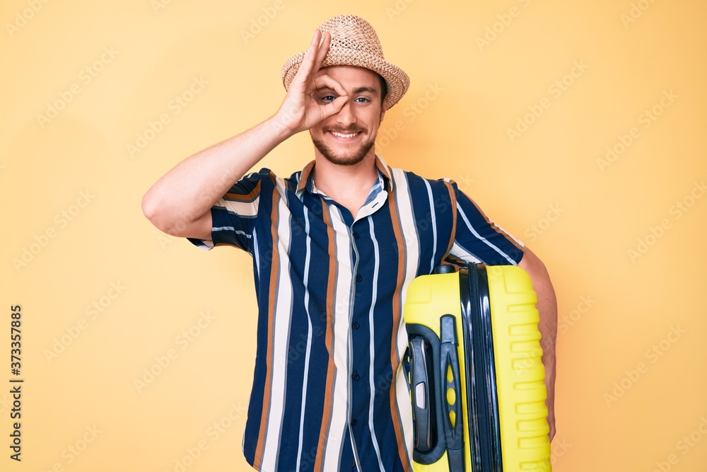 Young handsome man wearing summer hat holding cabin bag smiling happy doing ok sign with hand on eye looking through fingers Young handsome man wearing summer hat holding cabin bag smiling happy doing ok sign with hand on eye looking through fingers