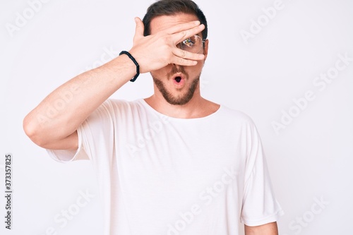 Young handsome man wearing casual clothes and glasses peeking in shock covering face and eyes with hand, looking through fingers afraid