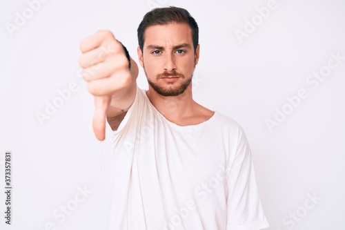Young handsome man wearing casual white tshirt looking unhappy and angry showing rejection and negative with thumbs down gesture. bad expression.