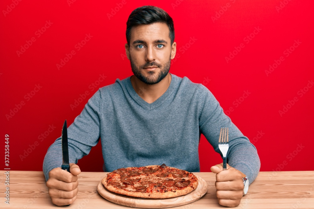 Handsome hispanic man eating tasty pepperoni pizza relaxed with serious expression on face. simple and natural looking at the camera.
