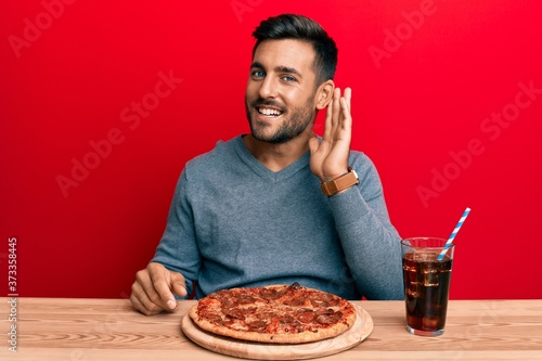 Handsome hispanic man eating tasty pepperoni pizza smiling with hand over ear listening and hearing to rumor or gossip. deafness concept.