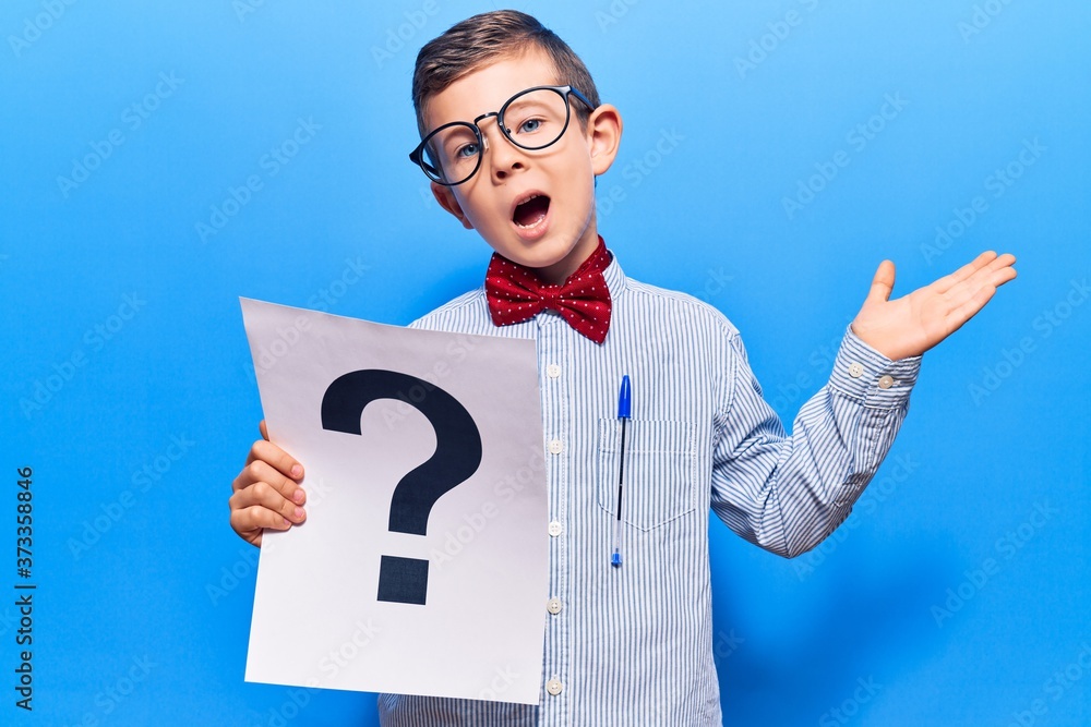 Cute blond kid wearing nerd bow tie and glasses holding question mark celebrating achievement with happy smile and winner expression with raised hand Cute blond kid wearing nerd bow tie and glasses holding question mark celebrating achievement with happy smile and winner expression with raised hand