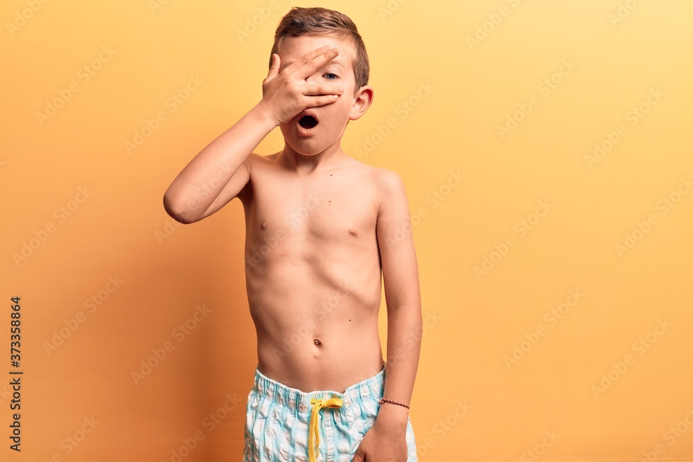 Cute blond kid wearing swimwear peeking in shock covering face and eyes with hand, looking through fingers afraid