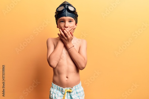 Cute blond kid wearing swimwear and swimmer glasses shocked covering mouth with hands for mistake. secret concept. © Krakenimages.com