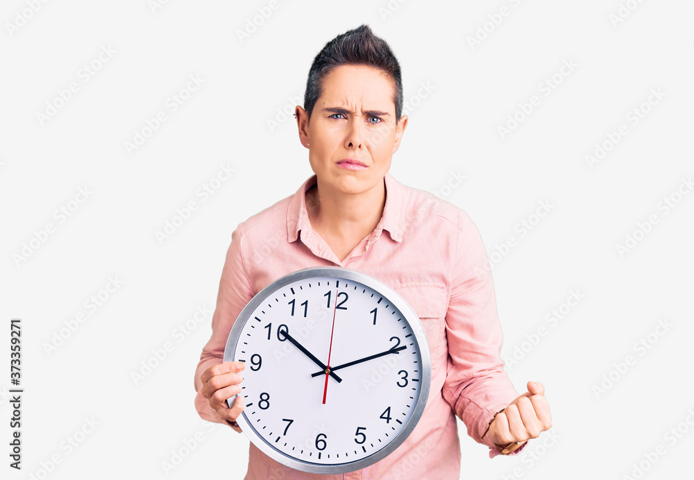 Young woman with short hair holding big clock annoyed and frustrated shouting with anger, yelling crazy with anger and hand raised Young woman with short hair holding big clock annoyed and frustrated shouting with anger, yelling crazy with anger and hand raised