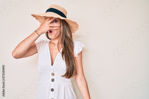 Young blonde girl wearing summer hat peeking in shock covering face and eyes with hand, looking through fingers afraid
