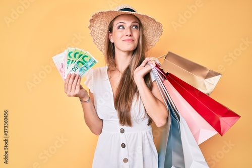 Young blonde girl holding shopping bags and israeli shekels smiling looking to the side and staring away thinking.