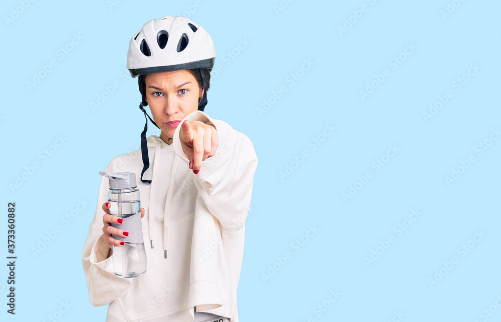 Beautiful brunette young woman wearing bike helmet and holding water bottle pointing with finger to the camera and to you, confident gesture looking serious Beautiful brunette young woman wearing bike helmet and holding water bottle pointing with finger to the camera and to you, confident gesture looking serious