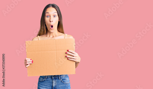 Beautiful brunette young woman holding cardboard banner with blank space scared and amazed with open mouth for surprise, disbelief face