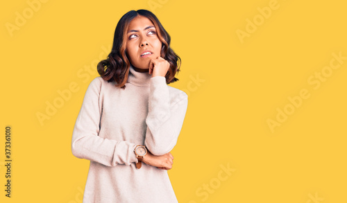 Young beautiful mixed race woman wearing winter turtleneck sweater with hand on chin thinking about question, pensive expression. smiling and thoughtful face. doubt concept.