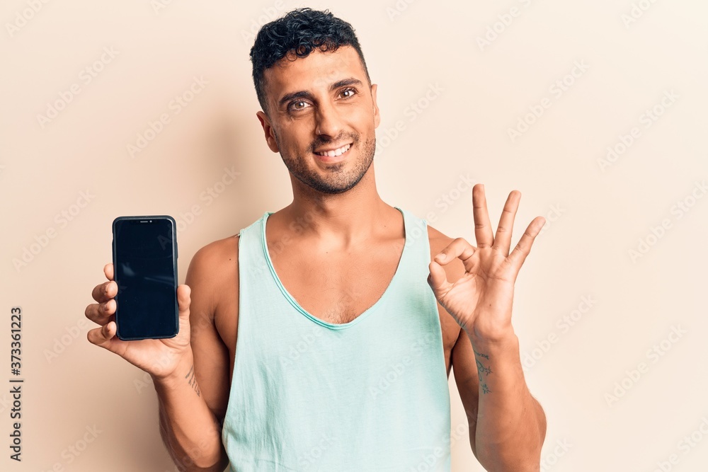 Young hispanic man holding smartphone showing screen doing ok sign with fingers, smiling friendly gesturing excellent symbol Young hispanic man holding smartphone showing screen doing ok sign with fingers, smiling friendly gesturing excellent symbol