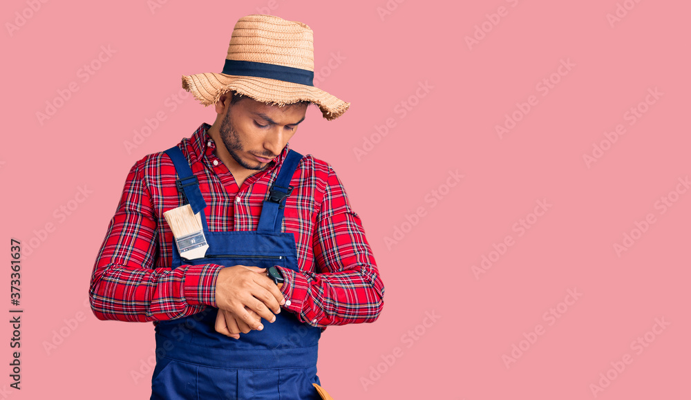 Handsome latin american young man weaing handyman uniform checking the time on wrist watch, relaxed and confident Handsome latin american young man weaing handyman uniform checking the time on wrist watch, relaxed and confident