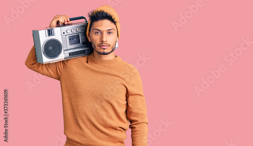 Handsome latin american young man holding boombox, listening to music relaxed with serious expression on face. simple and natural looking at the camera.