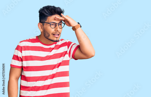 Handsome latin american young man wearing casual clothes and glasses very happy and smiling looking far away with hand over head. searching concept.