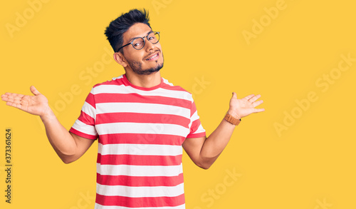 Handsome latin american young man wearing casual clothes and glasses clueless and confused expression with arms and hands raised. doubt concept.