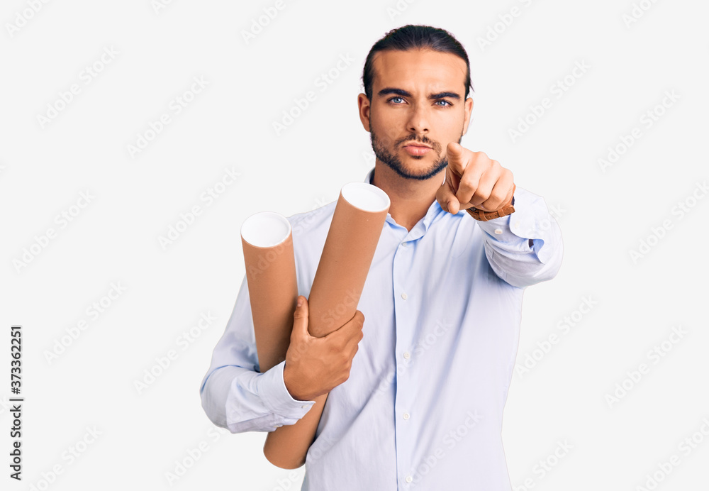 Young handsome man holding paper blueprints pointing with finger to the camera and to you, confident gesture looking serious Young handsome man holding paper blueprints pointing with finger to the camera and to you, confident gesture looking serious