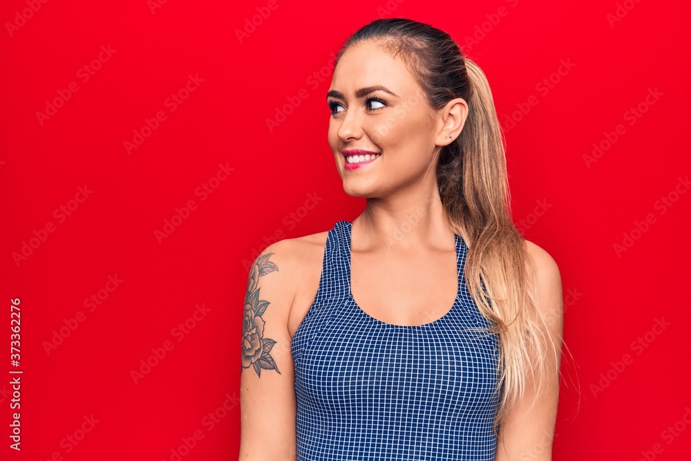 Young beautiful blonde woman wearing sporty t-shirt standing over isolated red background looking to side, relax profile pose with natural face and confident smile. Young beautiful blonde woman wearing sporty t-shirt standing over isolated red background looking to side, relax profile pose with natural face and confident smile.