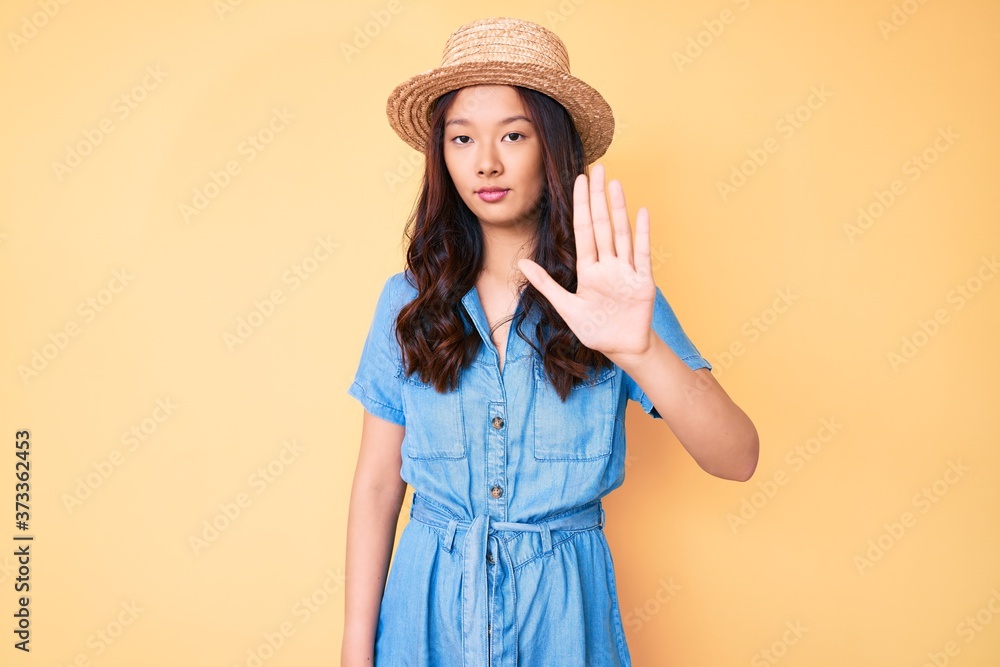 Young beautiful chinese girl wearing summer hat doing stop sing with palm of the hand. warning expression with negative and serious gesture on the face. Young beautiful chinese girl wearing summer hat doing stop sing with palm of the hand. warning expression with negative and serious gesture on the face.