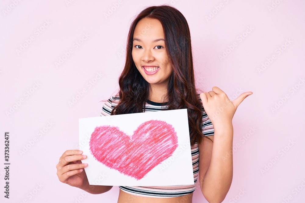 Young beautiful chinese girl holding heart shape paper pointing thumb up to the side smiling happy with open mouth Young beautiful chinese girl holding heart shape paper pointing thumb up to the side smiling happy with open mouth