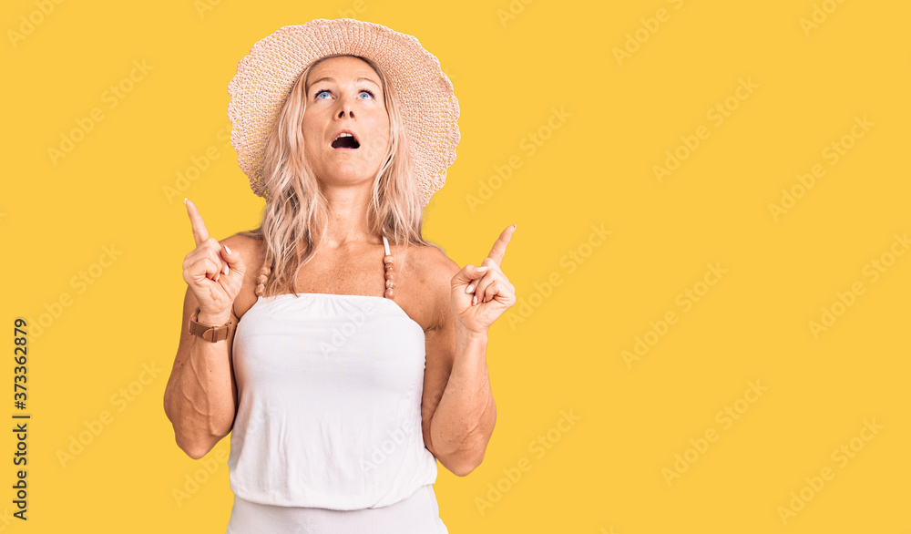Middle age fit blonde woman wearing summer hat amazed and surprised looking up and pointing with fingers and raised arms. Middle age fit blonde woman wearing summer hat amazed and surprised looking up and pointing with fingers and raised arms.
