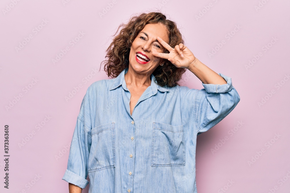 Middle age beautiful woman wearing casual denim shirt standing over pink background Doing peace symbol with fingers over face, smiling cheerful showing victory Middle age beautiful woman wearing casual denim shirt standing over pink background Doing peace symbol with fingers over face, smiling cheerful showing victory