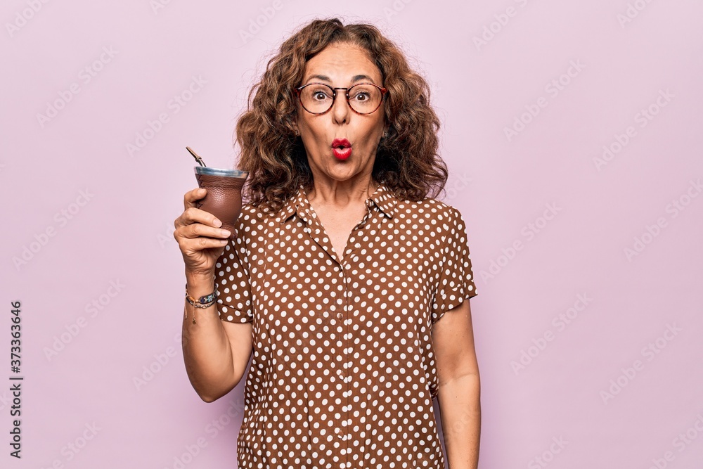 Middle age beautiful woman drinking cup of mate over isolated pink background scared and amazed with open mouth for surprise, disbelief face - obrazy, fototapety, plakaty 