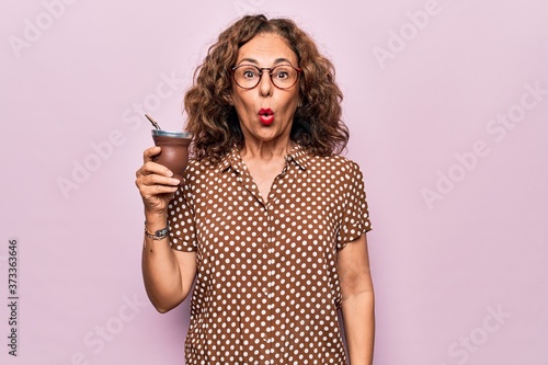 Middle age beautiful woman drinking cup of mate over isolated pink background scared and amazed with open mouth for surprise, disbelief face