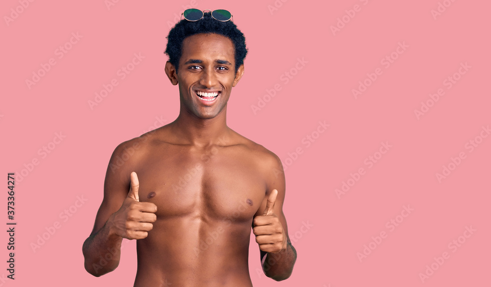 Fototapeta premium African handsome man wearing swimsuit and sunglasses success sign doing positive gesture with hand, thumbs up smiling and happy. cheerful expression and winner gesture.