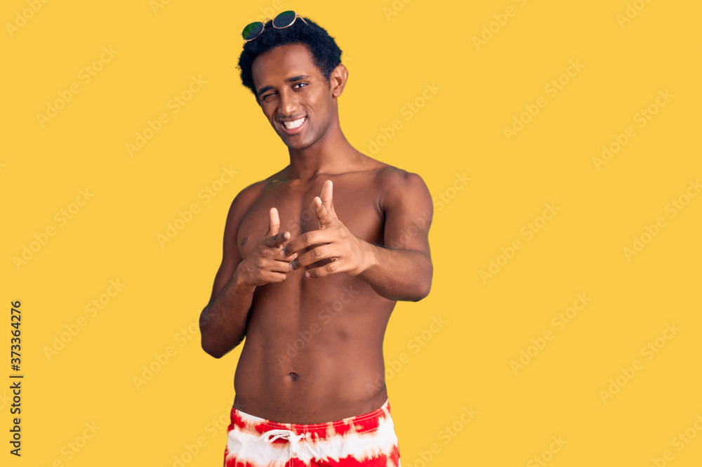 African handsome man wearing swimsuit and sunglasses pointing fingers to camera with happy and funny face. good energy and vibes.