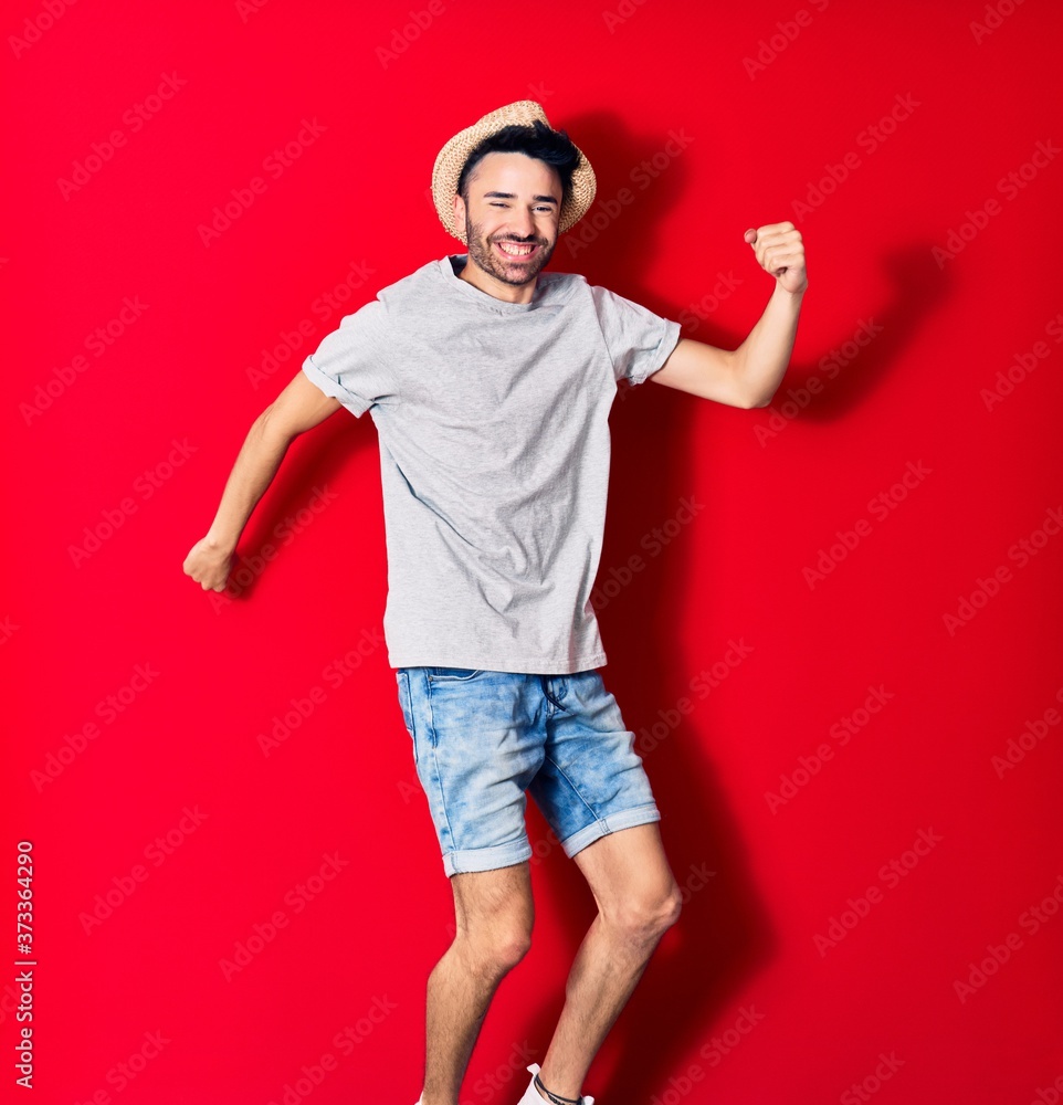 Young handsome hispanic man wearing casual clothes and summer hat smiling happy. Jumping with smile on face celebrating with fists up over isolated red background. Young handsome hispanic man wearing casual clothes and summer hat smiling happy. Jumping with smile on face celebrating with fists up over isolated red background.