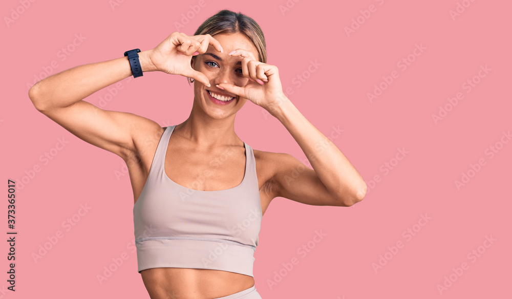 Beautiful caucasian woman wearing sportswear doing heart shape with hand and fingers smiling looking through sign Beautiful caucasian woman wearing sportswear doing heart shape with hand and fingers smiling looking through sign