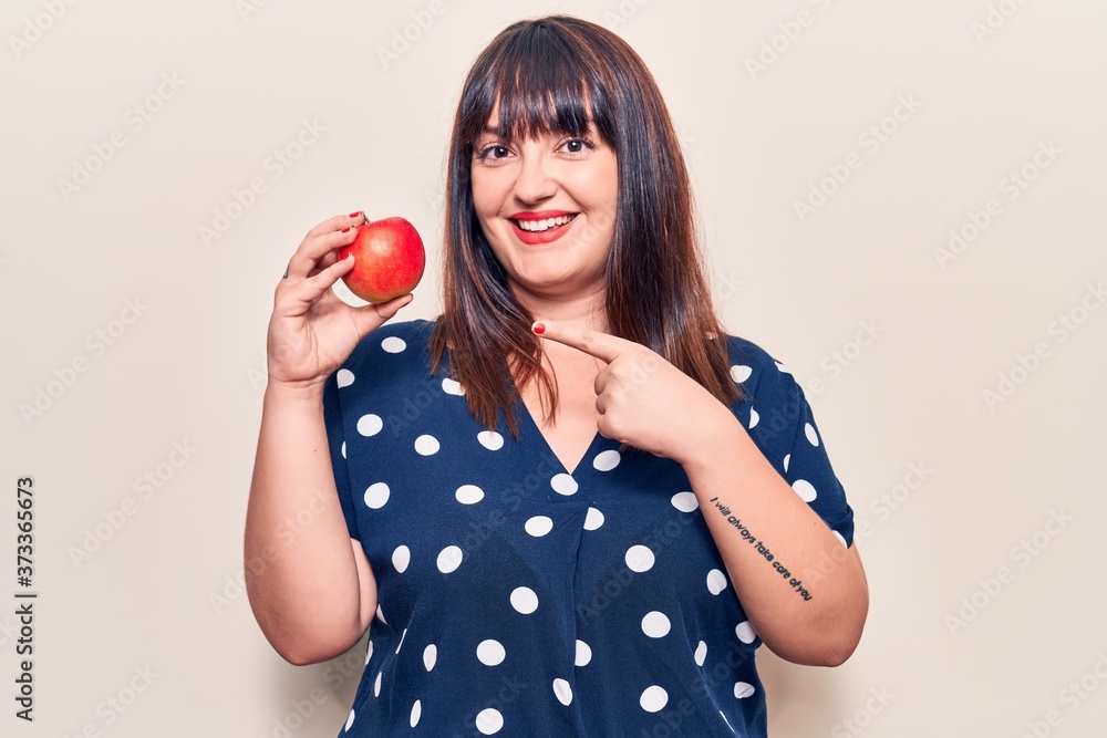 Young plus size woman holding red apple smiling happy pointing with hand and finger Young plus size woman holding red apple smiling happy pointing with hand and finger
