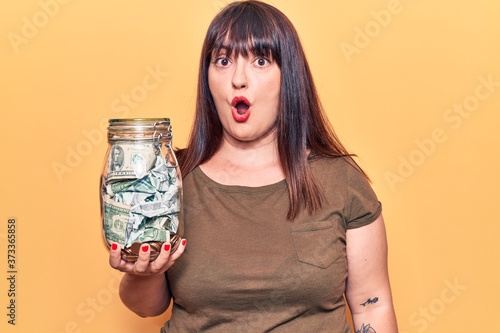 Foto Young plus size woman holding jar with savings scared and amazed with open mouth