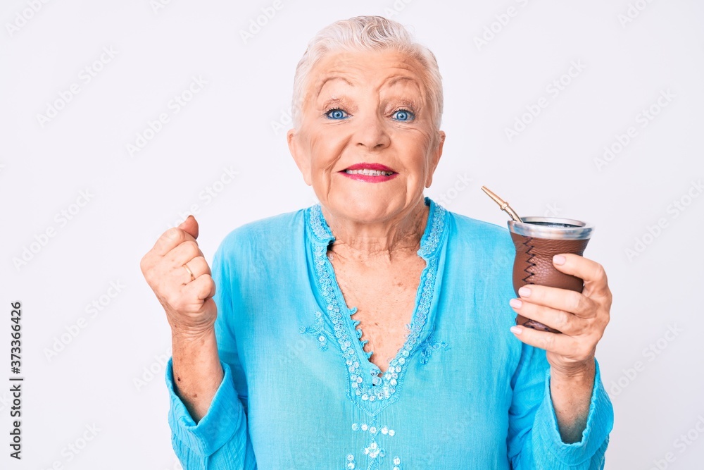 Senior beautiful woman with blue eyes and grey hair drinking mate infusion screaming proud, celebrating victory and success very excited with raised arm Senior beautiful woman with blue eyes and grey hair drinking mate infusion screaming proud, celebrating victory and success very excited with raised arm