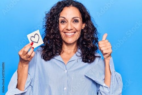 Middle age beautiful woman holding heart reminder smiling happy and positive, thumb up doing excellent and approval sign © Krakenimages.com