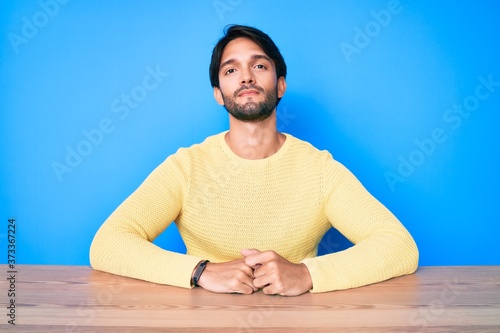 Handsome hispanic man wearing casual sweater sitting on the table with serious expression on face. simple and natural looking at the camera.