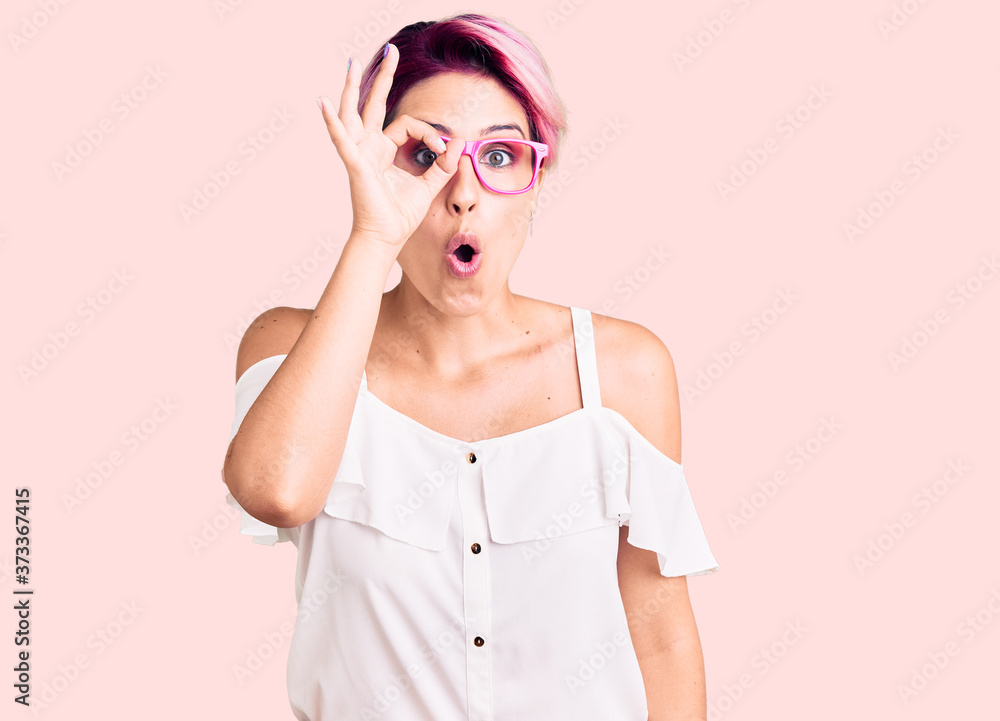 Young beautiful woman with pink hair wearing casual clothes and glasses doing ok gesture shocked with surprised face, eye looking through fingers. unbelieving expression. Young beautiful woman with pink hair wearing casual clothes and glasses doing ok gesture shocked with surprised face, eye looking through fingers. unbelieving expression.