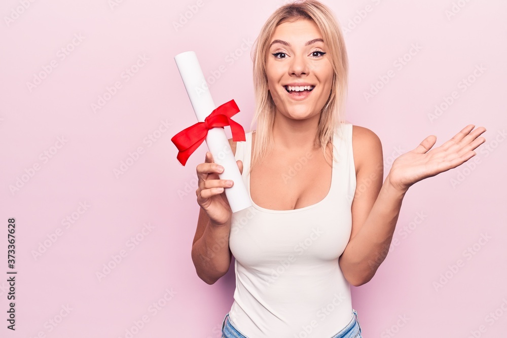 Young beautiful blonde student woman holding graduated degree diploma over pink background celebrating achievement with happy smile and winner expression with raised hand Young beautiful blonde student woman holding graduated degree diploma over pink background celebrating achievement with happy smile and winner expression with raised hand