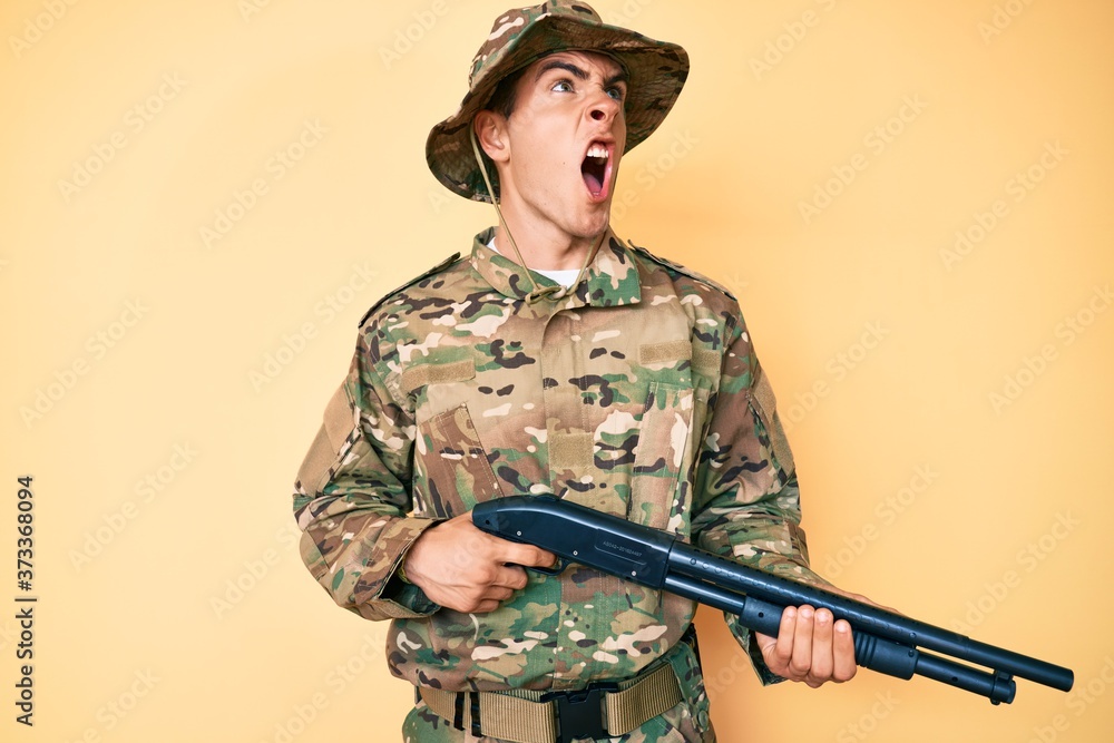 Young handsome man wearing camouflage army uniform holding shotgun angry and mad screaming frustrated and furious, shouting with anger. rage and aggressive concept. Young handsome man wearing camouflage army uniform holding shotgun angry and mad screaming frustrated and furious, shouting with anger. rage and aggressive concept.