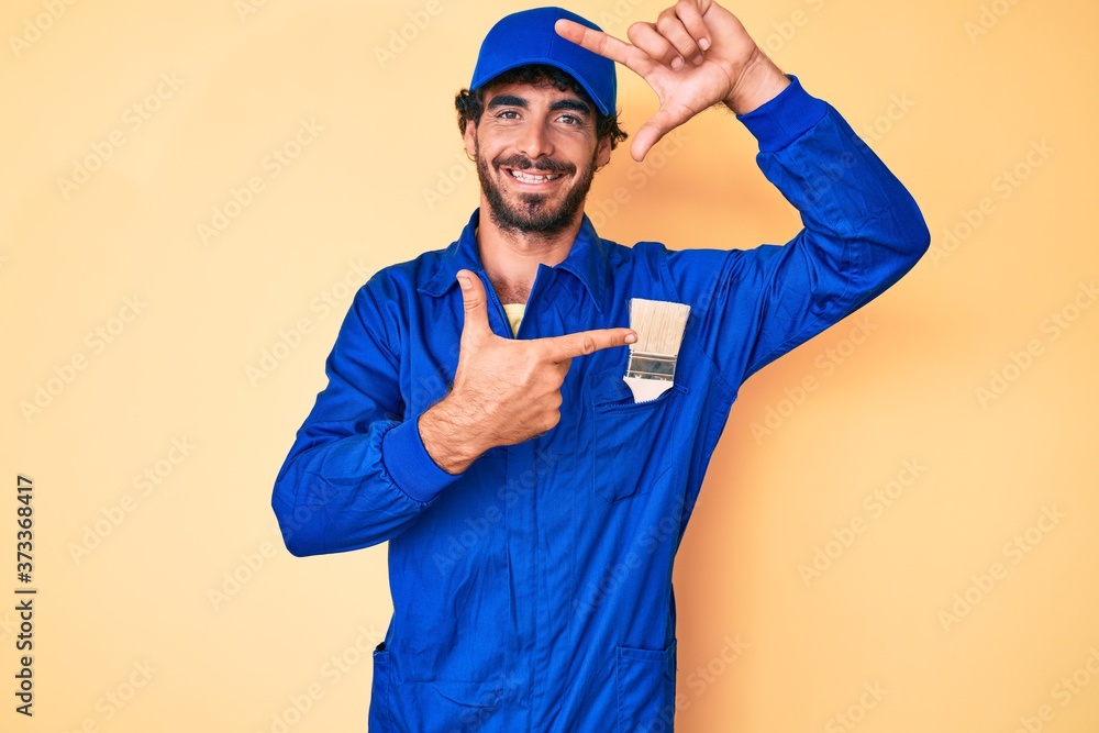 Handsome young man with curly hair and bear wearing builder jumpsuit uniform smiling making frame with hands and fingers with happy face. creativity and photography concept.