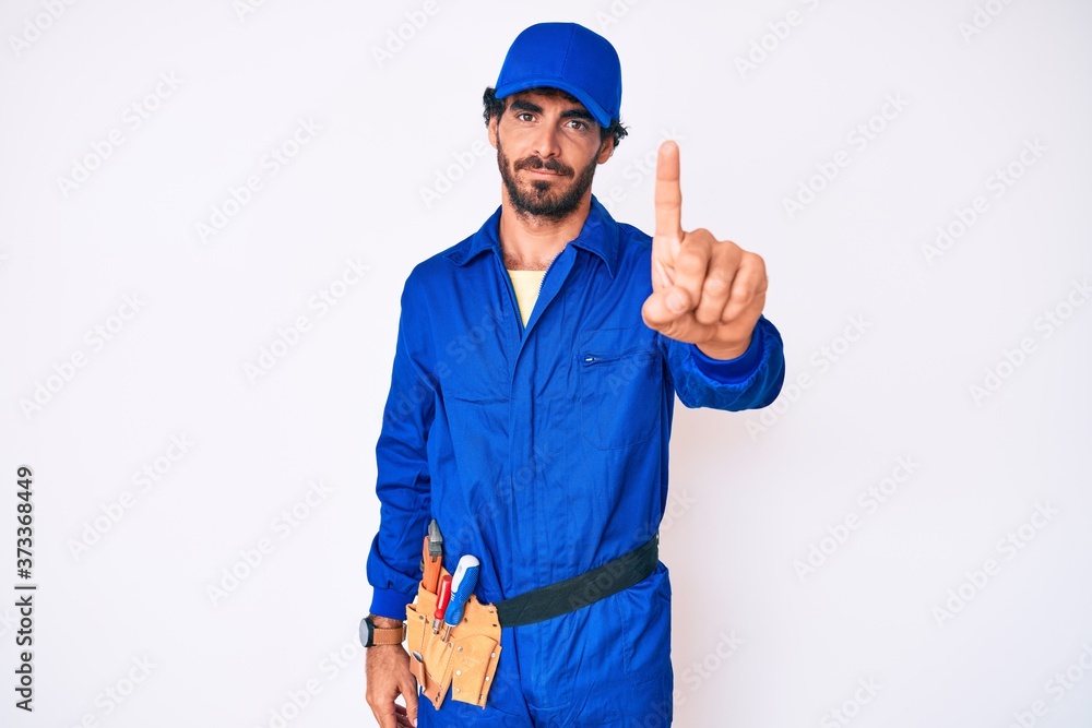 Handsome young man with curly hair and bear weaing handyman uniform pointing with finger up and angry expression, showing no gesture